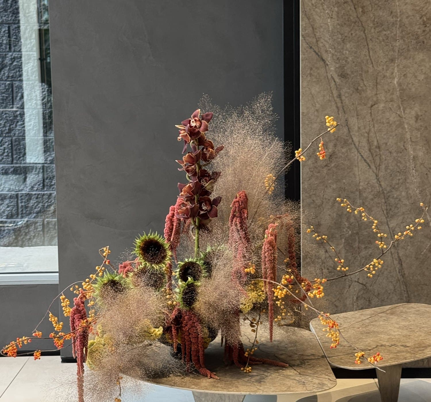 Sculptural Bloomiér arrangement of cymbidium orchids, amaranthus, sunflowers and smokebush on a stone table in a Vancouver showroom